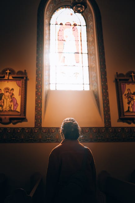 A person gazes at a sunlit stained glass window inside a church, evoking a sense of contemplation.